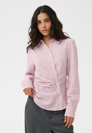 Femme aux longs cheveux foncés portant une chemise à carreaux rose boutonnée et un pantalon gris, debout devant un fond clair uni.