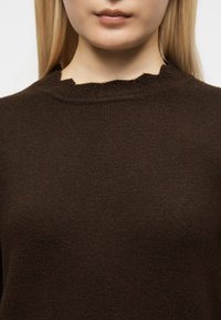 Gros plan d'une personne portant un haut en tricot texturé marron foncé avec un col rond légèrement froncé, ne montrant que les lèvres et le cou.