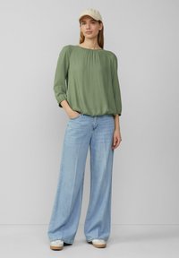 Olivgrüne Bluse mit rundem Ausschnitt, gerafften Ärmelbündchen, kombiniert mit hellblauen weiten Jeans und beigen Sneakers.