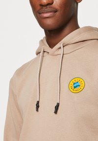 Beige hoodie i mjuk tyg med dragsko på huvan, med en rund patch med färgglada logodetaljer framtill.