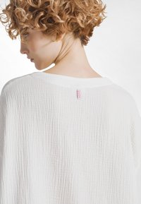 Witte top van gestructureerde stof met een los model, voorzien van een brede hals en een klein roze label aan de achterkant. Zacht en licht materiaal.