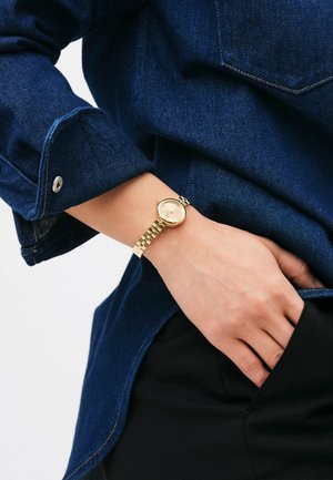 Montre dorée avec un cadran rond et minimaliste et un bracelet en métal maillé, portée au poignet à côté d'une chemise en denim bleu foncé.
