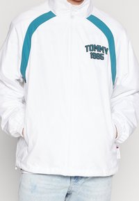 Bílá zipová bunda s tyrkysovými ramenními pruhy, předními kapsami a výšivkou "TOMMY 1985" na hrudi, nošena s světle modrými džíny.