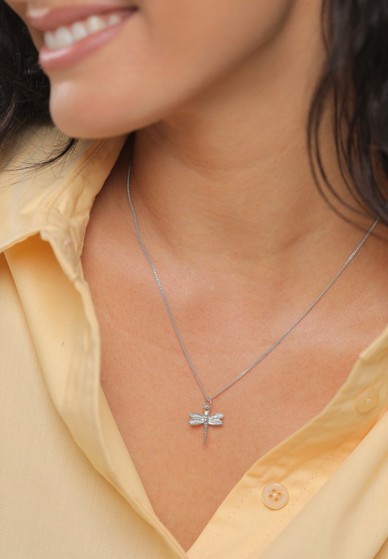 Strieborný prívesok v tvare vážky na jemnom retiazku, nosený s bledou žltou blúzkou s golierom, zdôrazňujúci štruktúrovaný dizajn a leštený povrch.