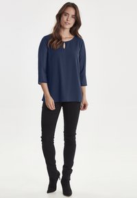 Blusa blu navy con scollo rotondo e dettagli a chiave, con maniche a tre quarti e fit morbido, abbinata a pantaloni neri slim.