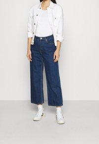 Biała dżinsowa kurtka na białej bluzce, zestawiona z luźnymi ciemnoniebieskimi dżinsami i białymi trampkami z czarnymi akcentami. Gładki materiał, swobodny krój.