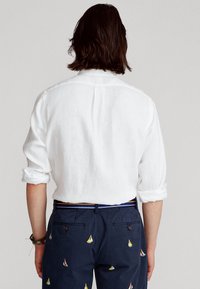 Vit linneskjorta med uppvikta ärmar, kombinerad med marinblå byxor med färgglad segelbådsbroderi. Synlig midjeband med ränder.