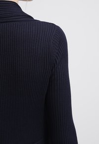 Mörkblå ribbad långärmad tröja som bärs av person, som visar ryggen och högra axeln mot en enfärgad ljus bakgrund.