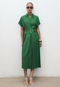 Green, knee-length shirt dress with short sleeves, a belted waist, and front pleats. Worn with white strappy sandals and black sunglasses.