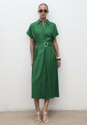 Green, knee-length shirt dress with short sleeves, a belted waist, and front pleats. Worn with white strappy sandals and black sunglasses.