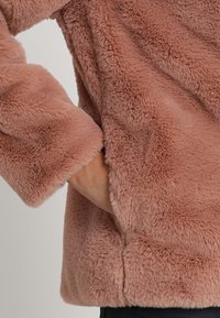 Faux furjacka i mjuk, puderrosa färg. Har en texturerad, fluffig yta och en avslappnad passform med en något kortad längd och breda ärmar.