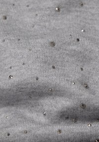 Grau gestrickter Stoff mit weicher Textur, versehen mit verstreuten metallischen Nieten verschiedener Größen auf der Oberfläche.