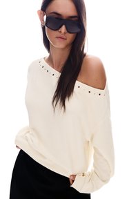 Pulover brez naramnic v kremni barvi z okrasnimi srebrnimi zakovicami ob izrezu. Mehka tekstura, sproščen kroj, v kombinaciji s črnimi hlačami in oglatimi sončnimi očali.