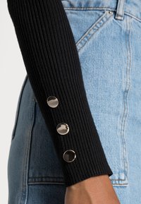 Haut noir à manches longues côtelé avec trois boutons métalliques au poignet, associé à un denim bleu. La texture est douce et ajustée.