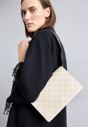 Beige patterned shoulder bag with a structured shape, featuring a brown textured strap. Worn on a model against a gray background.