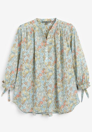 Frau trägt eine locker sitzende Bluse mit blauem, gelbem und orangefarbenem Blumenmuster und weiße Hose vor neutralem Hintergrund.