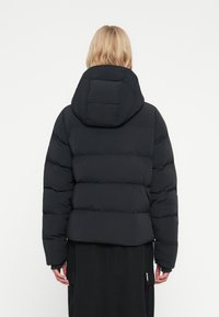 Blond person som har på sig en svart puffjacka med huva och en svart kjol, står vänd bort från en neutral vit bakgrund.