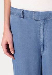 Svijetloplave traperice ravnog kroja. Imaju struk s petljama za remen i prošivene detalje sprijeda.