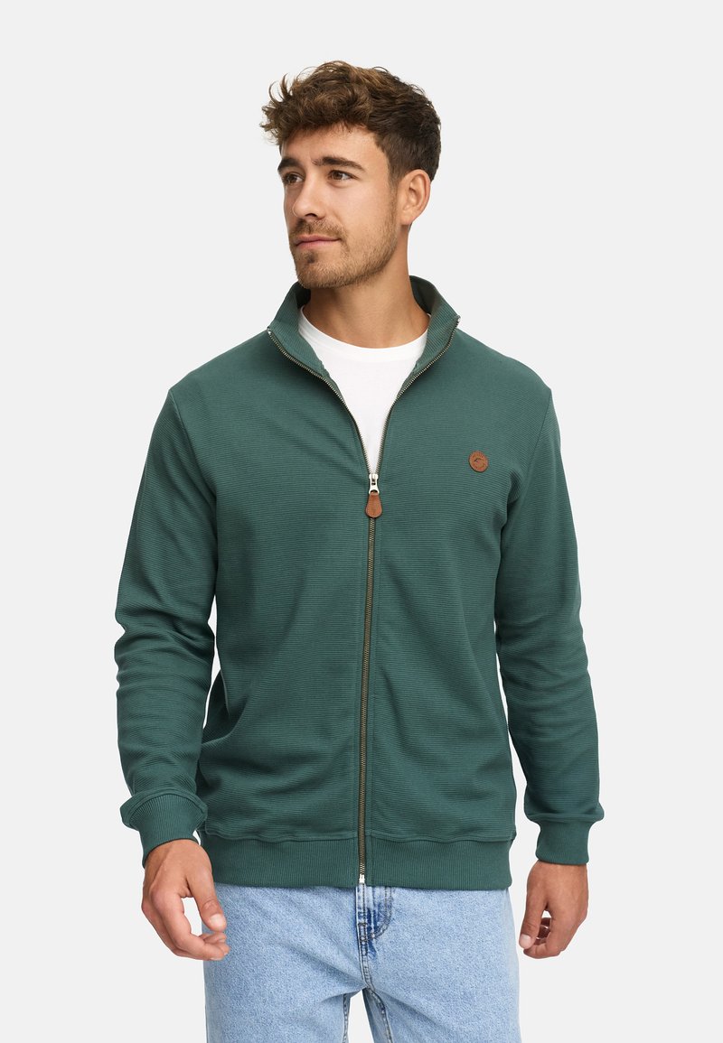 Ung mand iført en grøn zip-up jakke over en hvid t-shirt og lyseblå jeans, ser lidt til siden.