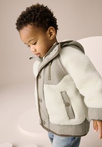 Veste grise et crème avec un devant en polaire douce et texturée, une fermeture éclair et des poches à boutons-pression. Dotée d'une capuche.