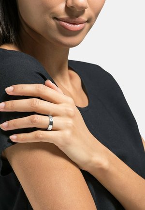 Donna con una maglietta nera che si tocca la spalla, indossa un anello d'argento sull'anulare, con un lucidalabbra rosa chiaro e unghie naturali.