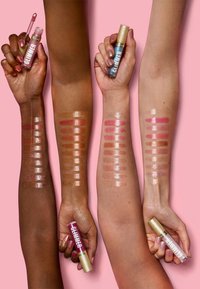 Tre arme viser lipgloss swatches på forskellige hudtoner. Glossene i rør har guldæsker med "GLIMMER" etiketten, sat op mod en rosa baggrund.