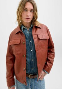 Brun læderjakke med to frontlommer, trykknapper og en let tætsiddende design, lagdelt over en denimskjorte.