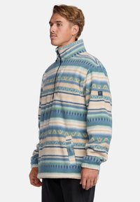 Fleece pulóver s vysokým golierom, ktorý má geometrický vzor v modrých, krémových a zelených odtieňoch. Obsahuje zips a predné vrecko.