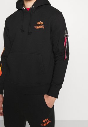Persoon gekleed in een zwart hoodie en broek met oranje Alpha Industries- en Hot Wheels-logo's, hand in de zak, rode ritssluitingdetail aan de mouw.