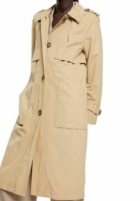 Trench-coat long beige avec grandes poches avant, fermeture par boutons, épaulettes et sangles de poignet réglables porté par une personne.