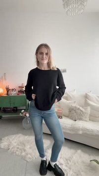 Žena plave kose stoji sa rukama u džepovima noseći crni džemper, plave traperice, bijele čarape i crne cipele u udobnoj dnevnoj sobi.