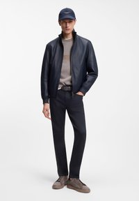 Ναυτική δερμάτινη τζάκετ, γκρι πουλόβερ με λογότυπο, μαύρη ζώνη, σκούρο παντελόνι και καφέ αθλητικά παπούτσια. Λεπτή υφή, καθαρός σχεδιασμός, ελάχιστες λεπτομέρειες.