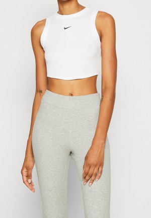 Henkilö, jolla on valkoinen hihaton lyhyt paita, jossa on Nike-logo, ja vaaleanharmaat korkeavyötäröiset leggingsit, seisoo yksivärisen taustan edessä.