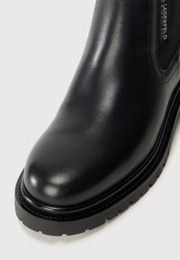 Black leather ankle boot with a smooth, glossy finish, rounded toe, thick black sole, and subtle stitching detail near the edge.