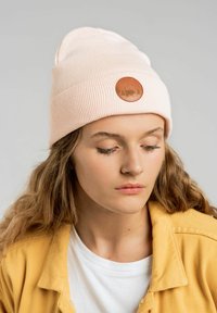 Jasnoróżowa, ryfrowana czapka beanie z okrągłą, tanową łatą ze wzorem przyrody. Nosi się ją przechyloną na głowie, widoczna miękka tekstura.