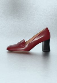 Mocassin femme en cuir rouge avec un bout carré, une bride décorative sur le dessus et un talon moyen noir et épais sur une surface lisse.