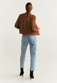 Veste en daim marron courte, jean taille haute bleu clair et bottines noires à talons. La vue de dos montre des cheveux tressés et un design simple.