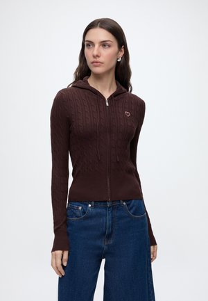 Kvinde iført en mørkebrun kabelstrikket lynlås-hættetrøje med en lille hjerteemblem og blå højtaljede jeans mod en ensfarvet hvid baggrund.