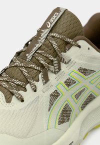 Beige und braun sportlicher Schuh mit strukturiertem Mesh-Obermaterial, gemusterten Schnürsenkeln, kontrastierenden gelben Akzenten und gepolsterter Sohlenkonstruktion.