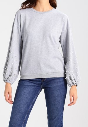 Felpa grigia con maniche lunghe e soffiate decorate con dettagli in perle bianche. Tessuto morbido, scollatura a girocollo e vita aderente. Indossata con jeans blu.