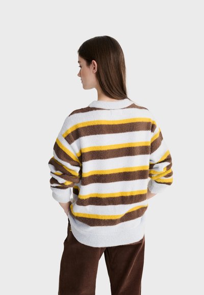 Femme aux longs cheveux bruns portant un pull blanc avec des rayures horizontales marron et jaune, associée à un pantalon marron, vue de profil.