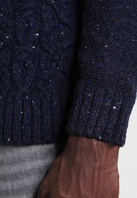 Granatowy sweter dzianinowy z teksturowanym wzorem w warkocze, prążkowanymi mankietami oraz wielokolorowymi drobinkami w tkaninie.