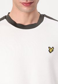 Vit bomull T-shirt med en mörkgrön ribbad rund hals och en liten gul broderad fågel-logotyp på vänster bröst.