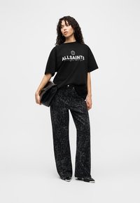 Μαύρο βαμβακερό t-shirt με γραφικό "ALLSAINTS", συνδυασμένο με παντελόνια φαρδιάς γραμμής με μαύρο λεοπάρ σχέδιο. Το μοντέλο κρατάει μια μαύρη τσάντα χειρός, φοράει μαύρες γόβες.