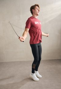 T-shirt rouge à manches courtes avec le texte "UFC", leggings noirs ajustés et baskets blanches. La personne fait de la corde à sauter dans un cadre intérieur.