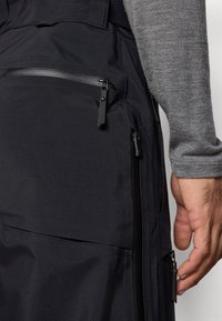 Μαύρο εξωτερικό παντελόνι με λείο, ματ ύφασμα, πολλαπλές τσέπες με φερμουάρ και κομψό σχεδιασμό. Διακοσμήσεις περιλαμβάνουν μαύρα φερμουάρ.