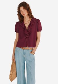 Blusa bordeaux con maniche a sbuffo e colletto arricciato, abbinata a jeans a gamba larga blu chiaro e una cintura beige strutturata.