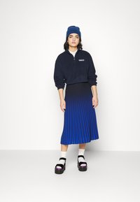 Ναυτικό κοντό φούτερ με φερμουάρ, μίντι φούστα μπλε με κλιμακωτές πτυχώσεις, μαύρες πλατφόρμες σανδάλια με μωβ λεπτομέρειες, λευκές κάλτσες.