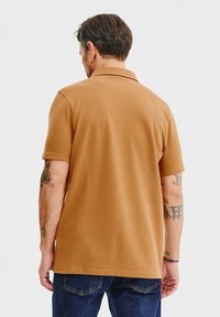 Brązowa koszulka polo z krótkim rękawem i klasycznym kołnierzykiem, o gładkiej fakturze i minimalnym zdobieniu, zestawiona z niebieskimi dżinsami.