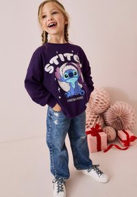 Lilla kapuutsiga sviiter, kus on "Stitch" graafika ja sädelevad aktsendid, koos helesiniste rebenditega teksade ja valgete tossudega.